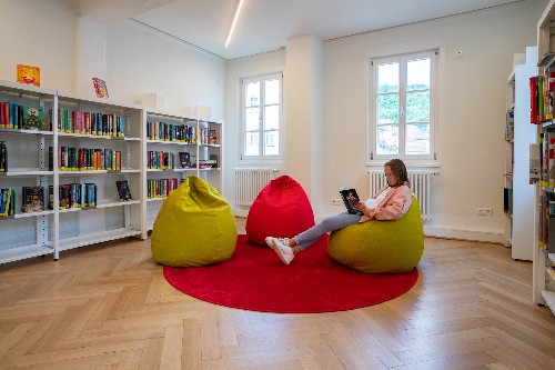 Blick in einen Raum in der Stadtbücherei, Bücheregale davor Sitzgruppe mit drei farbeigen Sesseln und einem lesenden Kind