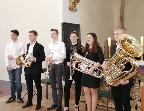 zwei Musikerinnen und vier Musiker stehen in Reihe nebeneinander, sie haben ihre Instrumente in der Hand: Tuba, Trompete, Horn