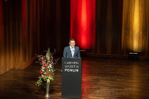 ein Herr am Rednerpult im Carmen Würth Forum, daneben ein großes Blumengebinde in rot-weiß