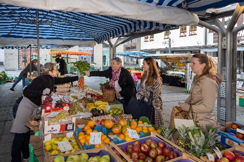 drei Frauen stehen an einem Obst- und Gemüsestand und werden von einer Verkäuferin bedient