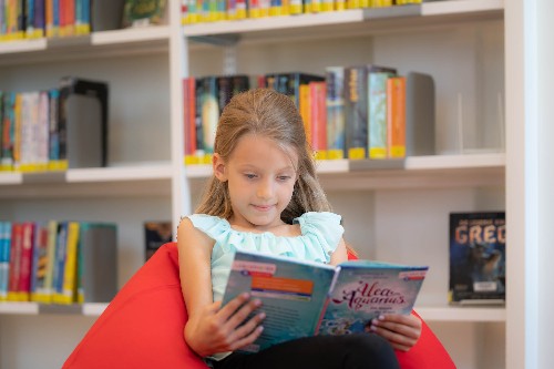 Ein Mädchen sitzt auf einem roten Sessel vor einem Bücherregal und schaut in ein Buch.