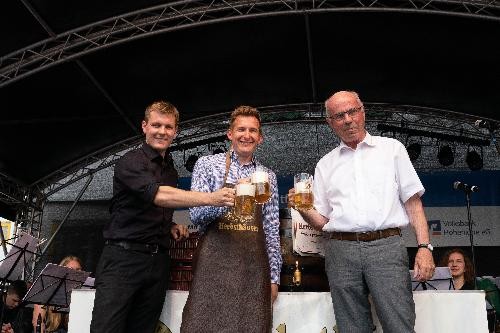 Drei Herren auf der Bühne mit einem vollen Bierkrug in der Hand