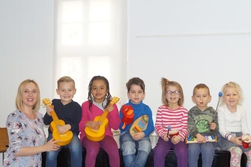 sechs Kinder sitzen nebeneinander, jedes hat ein kleines Musikinstrument in der Hand. Links steht die Lehrerin