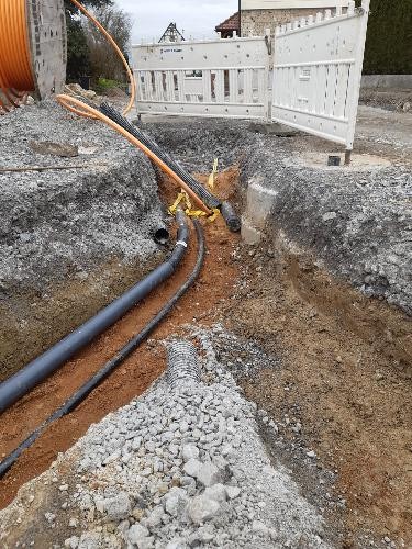 Blick in offene Baugrube in der ein schwarzes Rohr und ein schwarzes Kabel liegen, daneben steht eine große Holzrolle mit orangefarbenem Kabel