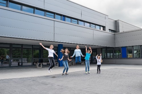 fünf Kinder springen freudig in die Luft, recken die Arme nach oben, im Hintergrund die Außenfassade des Ganerben-Gymnasiums
