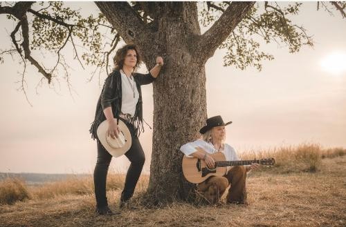 zwei Frauen in Cowboy-Kleidung. Eine lehnt sich an einen Baum, die andere sitzt am Baum mit einer Gitarre in der Hand