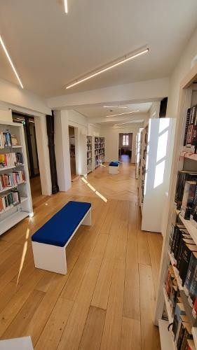 Blick in einen langen Bücherei-Raum, heller Holzboden, weiße Bücherregale und blaue Sitzbänke