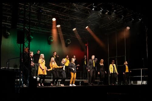 acht Personen in gelb und scharzer Kleidung singen auf einer Bühne
