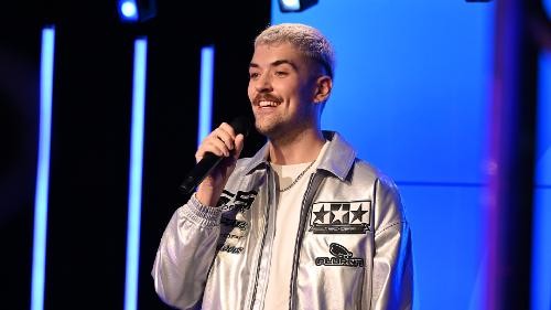 ein blonder Mann in silberfarbener Jacke hat ein Mikro in der Hand, der Hinterggrund it blau