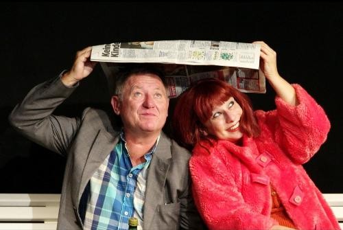Zwei Schauspieler, ein männlicher und eine weibliche, schützen sich vor REgen mit einer Zeitung über dem Kopf.