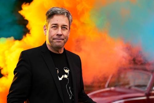 ein grauhaariger schwarz gekleider Mann, im HIntergrund lodernde Flammen