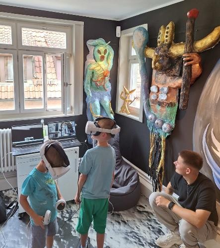 zwei Jungs haben VR-Brillen auf, im Hintergrund ein junger Mann in der Hocke an der Wand hängen bute Alienfiguren