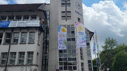 vier bunte Fahnen wegen an den Fahnenmasten vor der der weißen Rathausfassade im Wind