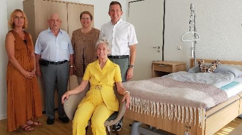 zwei Damen und zwei Herren stehen nebeneinander, davor sitzt einen Dame ganz in gelb gekleidet. Rechts im Bild ist ein Pflegebett zu sehen