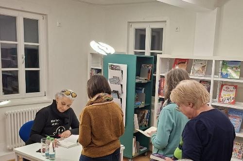 Die Autorin sitzt in der Stadtbücherei auf einem Stuhl mit einem Tisch vor hr und gibt unterschriten. Die Leute stehen in der Schlange.