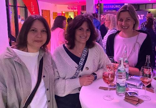 Drei Frauen stehen vor einem weißen stehtisch und haben gute Laune. Im Hintergrund viele Menschen und buntes Licht.