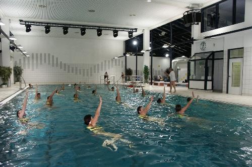 Blick ins große Wasserbecken in dem mehrere Personen Gymnastik machen, Arme hoch, Anleitung von Trainerin am Beckenrand