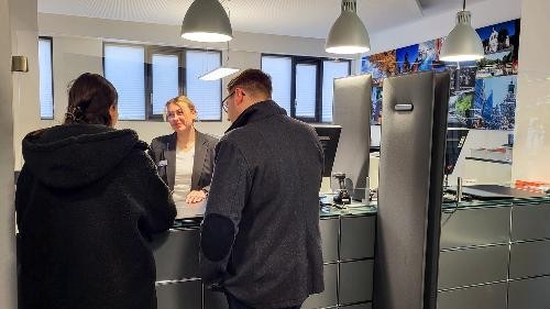 Eine Dame und ein Herr stehen vor einer Theke - sie sind von hinten zu sehen und sprechen mit einer blonden Dame, die hinter der Theke steht