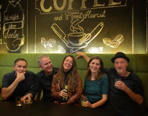 Fünf Personen, zwei Frauen und drei Männer sitzen in gemütlicher atmosphäre nebneinander. im Hintergrund eine Grüne Wand mit Kaffee bemalung.