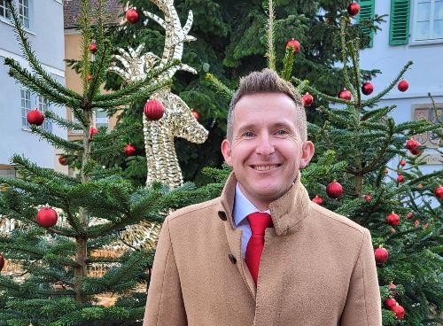Bürgermeister Stefan Neumann steht mit beigen Mantel und roter Krawatte lächelnd vor einem geschmücktem Weihanchtsbaum.