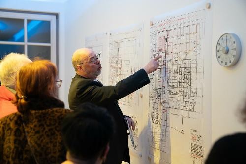 ein Herr zeigt auf einen Plan an der Wand, zwei Damen schauen ihm zu
