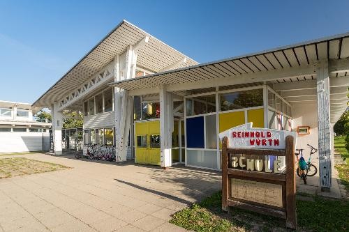 Ein schulgebäude aus Holz mit einer wei7gelben Fassade. Blauer Himmel.