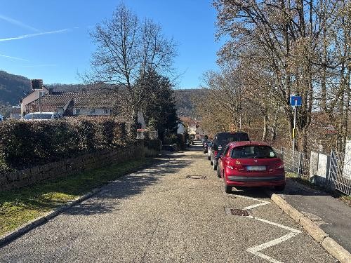 Auf dem Foto sieht man eine schmale Straße. Rechts ein Gehweg und links Hecken und Häuser.