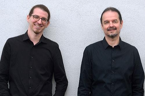 Zwei Männer stehen mit schwarzem Hemd vor einer weißen Wand und lächeln in die Kamera.