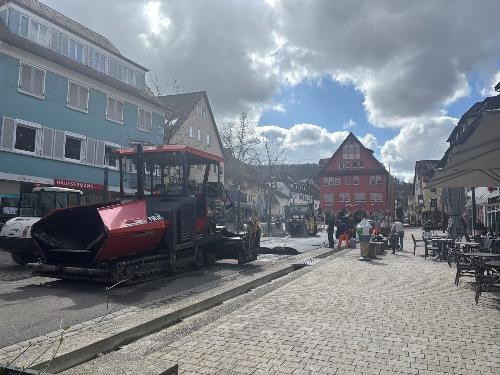 Blick auf die Baustelle: Eine rote Teermaschine im Vordergrund und dahinter eine gelbe Walze und Bauarbeiten, die den frisch aufgebrachten Teer verarbeiten