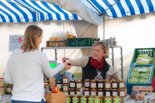Zwei Personen stehen an einem Marktstand und übergeben sich frisches Gemüse. Eine Person reicht der anderen eine Auswahl an Obst und Gemüse über den Verkaufstisch