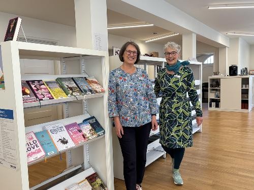 Zwei Frauen, Charlotte Sigrun Hellinger und Udine Löhlein, stehen nebeneinander in einer Bibliothek vor einem Regal mit zahlreichen Büchern. Sie befinden sich in der Stadtbücherei Künzelsau und blicken in die Kamera, während sie von Büchern umgeben sind.