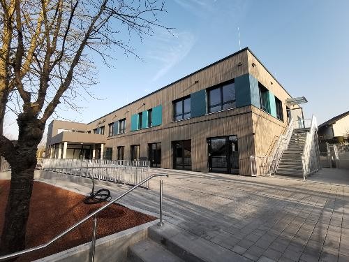 Außenansicht der neuen Grundschule in Amrichshausen mit moderner Holzfassade, klarer Architektur und großzügigen Fensterflächen, eingebettet in eine offene und freundliche Umgebung; die Öffentlichkeit ist zum Tag der offenen Tür eingeladen.