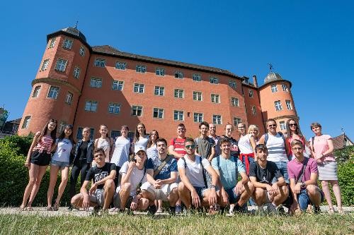 Eine Gruppe von Sprachschülerinnen und Sprachschülern steht vor einem Schloss. Im Hintergrund ist das historische Gebäude unter einem klaren, blauen Himmel zu sehen. Die Jugendlichen posieren gemeinsam im Freien und lächeln in die Kamera.