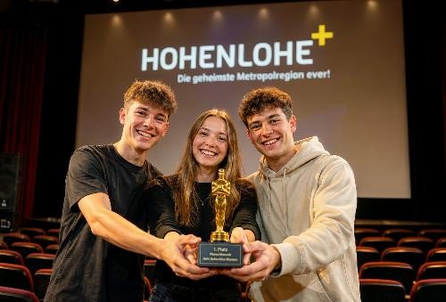 Auf einer Bühne in einem Kinosaal stehen drei junge Erwachsene dicht nebeneinander und lächeln in die Kamera. Gemeinsam halten sie eine goldene Siegerfigur mit der Aufschrift „1. Platz“. Im Hintergrund ist auf einer großen Leinwand das Logo „HOHENLOHE+“ mit dem Slogan „Die geheimste Metropolregion ever!“ zu sehen. Die Stimmung wirkt feierlich und stolz.