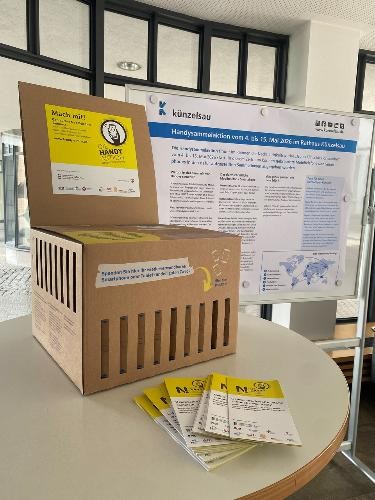 Eine Sammelbox aus Karton für ausgediente Mobiltelefone und Tablets steht auf einem Tisch im Rathaus Künzelsau. Daneben liegen Informationsflyer. Im Hintergrund informiert eine Ausstellungstafel über die Handysammelaktion im Rahmen der Nachhaltigkeitswoche „Gutes Klima für Hohenlohe“ und die Wiederverwendung wertvoller Rohstoffe.