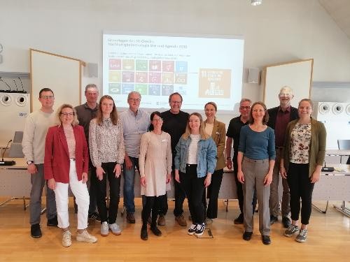 Gruppenfoto von Teilnehmenden eines Workshops in einem Tagungsraum. Die Personen stehen vor einer Präsentation zur Nachhaltigkeitsstrategie Baden-Württemberg und den globalen Nachhaltigkeitszielen. Der Workshop beschäftigte sich mit dem kommunalen Nachhaltigkeits-Check der Stadt Künzelsau.