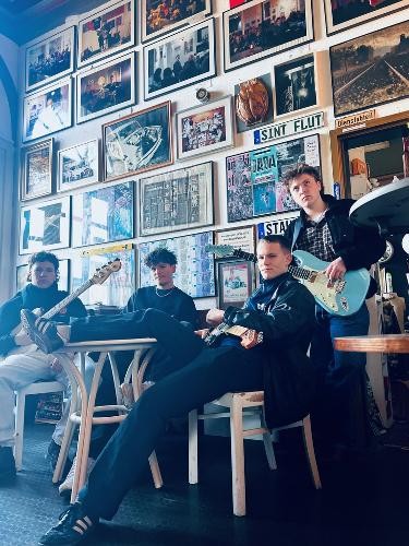 Vier junge Musiker der Rockband Spielzimmer posieren in einem gemütlichen, mit vielen gerahmten Bildern dekorierten Innenraum. Zwei Bandmitglieder sitzen an einem Tisch, einer lehnt entspannt auf einem Stuhl, während ein weiterer mit einer hellblauen E-Gitarre steht. Die Musiker tragen dunkle Freizeitkleidung und blicken selbstbewusst in die Kamera. Die Atmosphäre wirkt kreativ und ungezwungen und vermittelt den Eindruck einer jungen Band mit Bezug zum klassischen Rock.