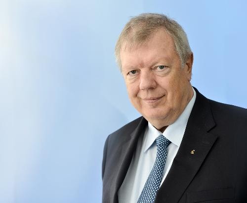 Porträt eines älteren Mannes mit kurzen grauen Haaren. Er trägt ein dunkles Sakko, ein hellblaues Hemd und eine hellblaue Krawatte mit kleinem Muster. Die Person blickt direkt in die Kamera und hat einen neutralen, leicht freundlichen Gesichtsausdruck. Der Hintergrund ist unscharf und in hellen Blau- und Grautönen gehalten. Die Aufnahme zeigt den Mann von der Brust aufwärts und wirkt wie ein professionelles Business- oder Pressefoto.