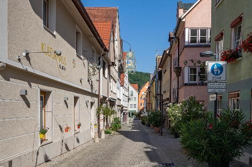 Eine schmale, gepflasterte Altstadtgasse in Künzelsau an einem sonnigen Tag. Links und rechts stehen mehrstöckige Häuser in hellen Pastellfarben mit roten Ziegeldächern. Vor den Gebäuden befinden sich zahlreiche Pflanzen und Blumenkästen. Über der Gasse hängen historische Wirtshausschilder. Im Hintergrund ist ein bewaldeter Hügel zu sehen. Die Straße ist nahezu menschenleer und vermittelt eine ruhige, historische Atmosphäre.