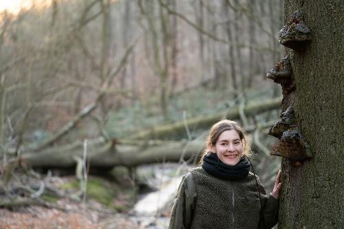 Die Naturfotografin Farina Graßmann steht in einem lichten Wald neben einem Baumstamm. Sie lächelt in die Kamera und legt eine Hand an den Stamm. An der Rinde wachsen mehrere dunkle Baumpilze übereinander. Im Hintergrund sind umgestürzte Bäume und ein kleiner Bach zu erkennen, die durch die geringe Schärfentiefe verschwommen wirken. Die Szene vermittelt eine ruhige, naturnahe Atmosphäre.