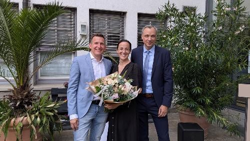 Drei Personen stehen vor dem Rathaus beziehungsweise einem Verwaltungsgebäude und blicken in die Kamera. Die Person in der Mitte hält einen Blumenstrauß. Links steht Bürgermeister Stefan Neumann in hellem Sakko, rechts ein Mann im dunklen Anzug. Im Hintergrund sind große Pflanzkübel mit Palmen und blühenden Sträuchern sowie die Fassade des Gebäudes zu sehen. Das Foto entstand anlässlich der Wiederwahl von Stefan Neumann zum Bürgermeister der Stadt Künzelsau am 14. Juni 2026.