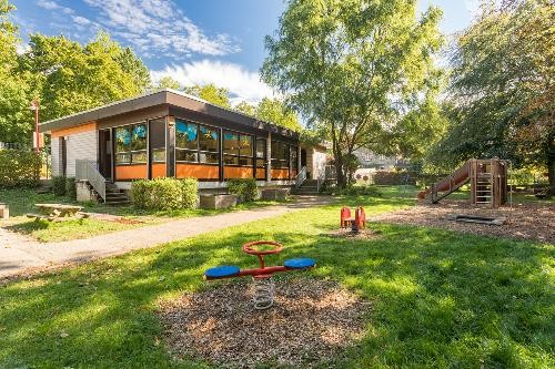 Zu sehen ist der Kindergarten Morsbach an einem sonnigen Tag. Das eingeschossige Gebäude mit Flachdach steht inmitten eines großen, grünen Gartens. Vor dem Haus befinden sich Spielgeräte, darunter eine Wippe auf einer Feder und ein Kletter- und Rutschgerüst. Hohe Bäume spenden Schatten und umgeben das Gelände. Große Fenster lassen viel Licht in die Räume. Die Anlage wirkt freundlich, naturnah und bietet viel Platz zum Spielen und Bewegen im Freien.
