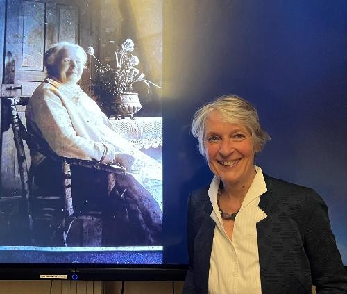 Eine Frau mit kurzen grauen Haaren und dunklem Blazer steht vor einem blauen Hintergrund neben einem großen Bildschirm. Auf dem Bildschirm ist ein historisches Foto von Karoline Breitinger zu sehen, die sitzend an einem Tisch dargestellt wird. Die Frau ist die Historikerin Cornelia Strauß, die über Breitingers Leben und Wirken referiert.