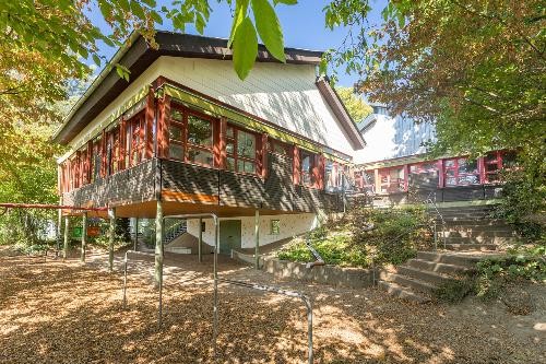 Außenansicht des Kindergartens Weckrain an einem sonnigen Tag. Das Gebäude mit großen Fensterfronten und dunkler Holzverkleidung steht erhöht auf Stützen inmitten eines grünen, von Bäumen umgebenen Außengeländes. Rechts führen Treppen zu den Eingängen, unter dem Gebäude befindet sich ein überdachter Spielbereich. Das Bild zeigt die naturnahe Lage und die großzügige Architektur der Einrichtun