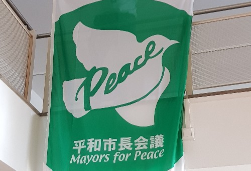 Bürgermeister Neumann hält eine weiß-grüne Flagge, mit Motiv Friedenstaube und Schriftzug "Bürgermeister für den Frieden"