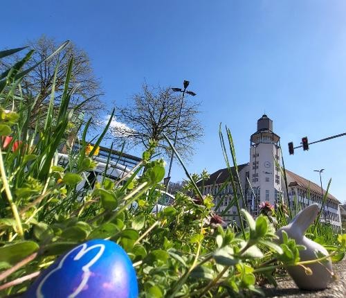 ein blaues Osterei liegt vorne im Foto im grünen Gras, rechts ist eine kleine beigefarbene  Hasenfigur und im Hintergrund das Rathaus mit dem Turm zu sehen