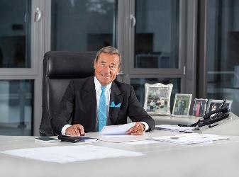 Mann in schwarzem Anzug und blauer Krawatte sitzt am Schreibtisch.