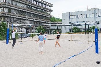 vier Personen spielen Beachvolleyball, im Hintergrund Hochschulgebäude