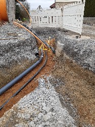 Blick in offene Baugrube in der ein schwarzes Rohr und ein schwarzes Kabel liegen, daneben steht eine große Holzrolle mit orangefarbenem Kabel