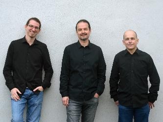 drei Herren mit schwarzen Hemden stehen nebeneinander vor einer hellgrauen Wand